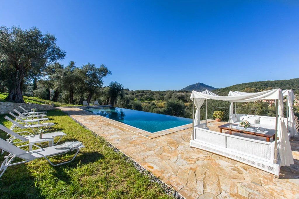 Infinity pool with white patio furniture, mountain views and olive trees in garden