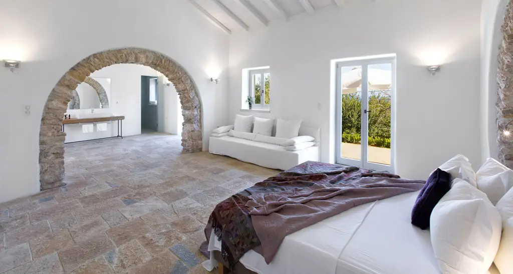 A villa bedroom showing a bed, white walls with a stone arch doorway and a white sofa.