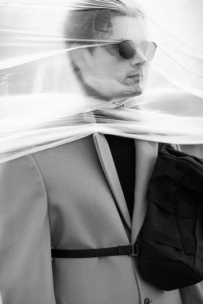 Model wearing a coat, sun glasses and bag with plastic sheeting covering face.