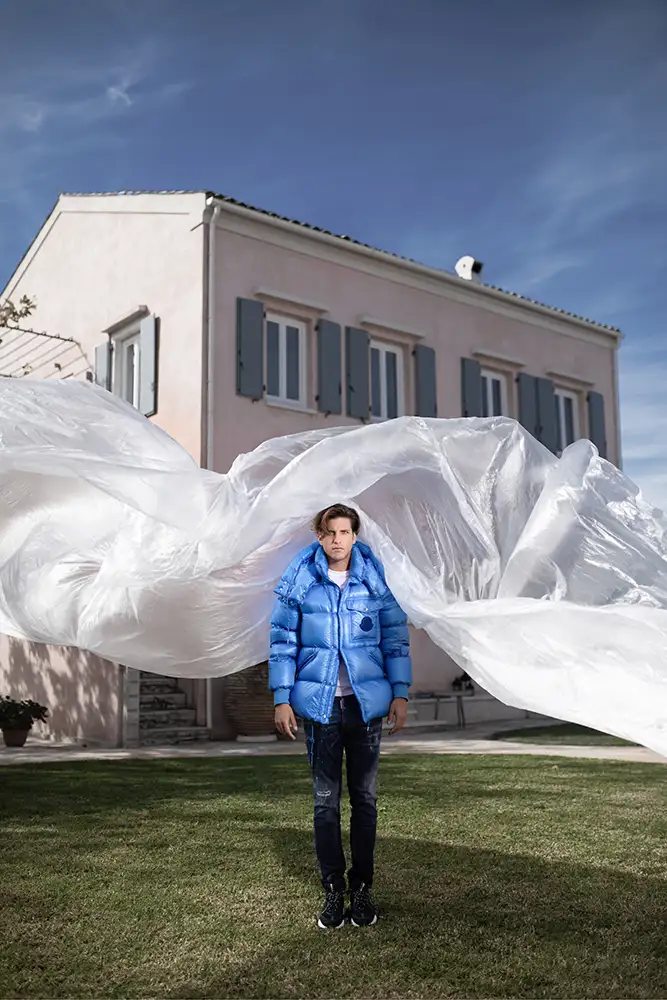Model wearing blue jacket in garden with plastic sheeting blowing around.