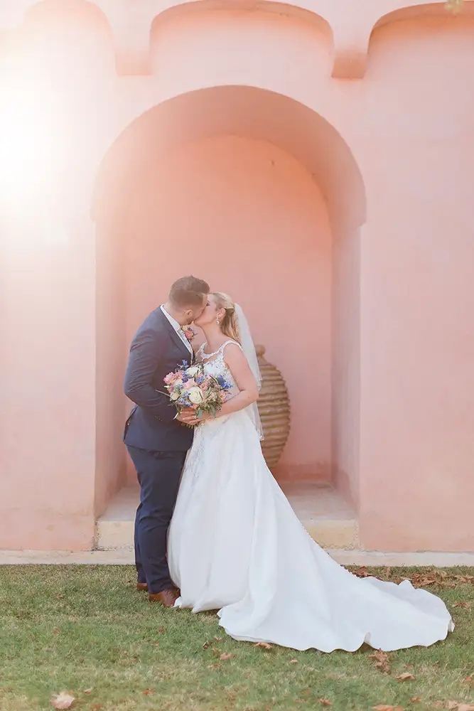 Bride and groom kissing with a peach coloured wall as a backdrop.