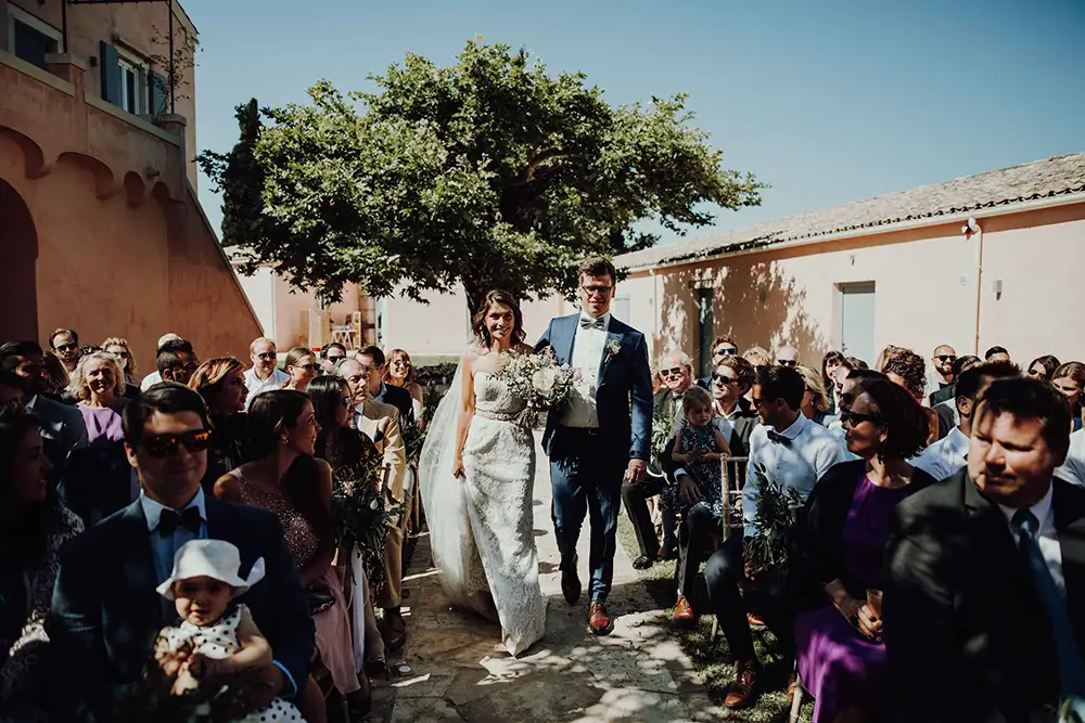 Bride walking down path inbetween guests with olive tree in background.