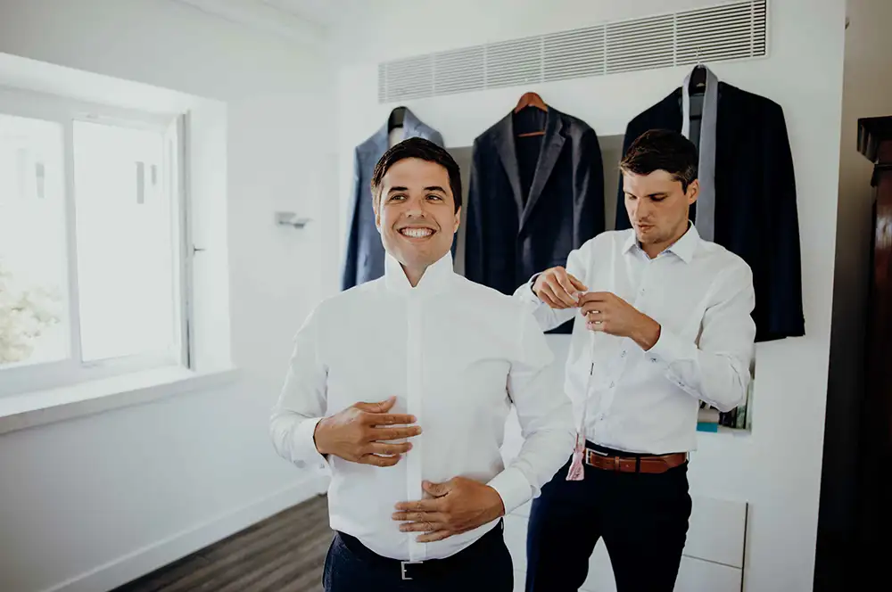 Groom and best man getting ready in the room with suits hanging up.