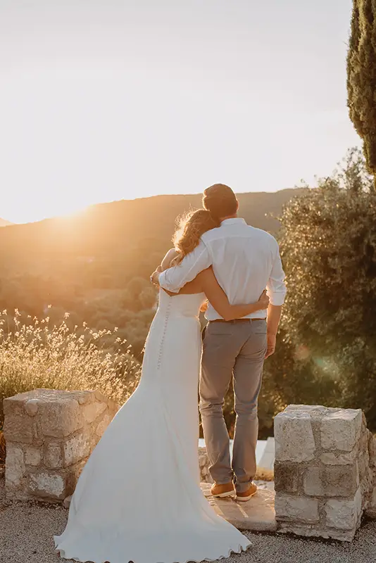 Bride and groom with arms around each other looking out as the sun sets behind a mountain