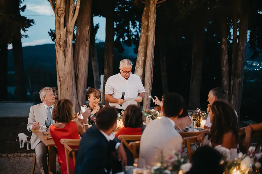 Wedding party sitting around table looking towards guest making speech